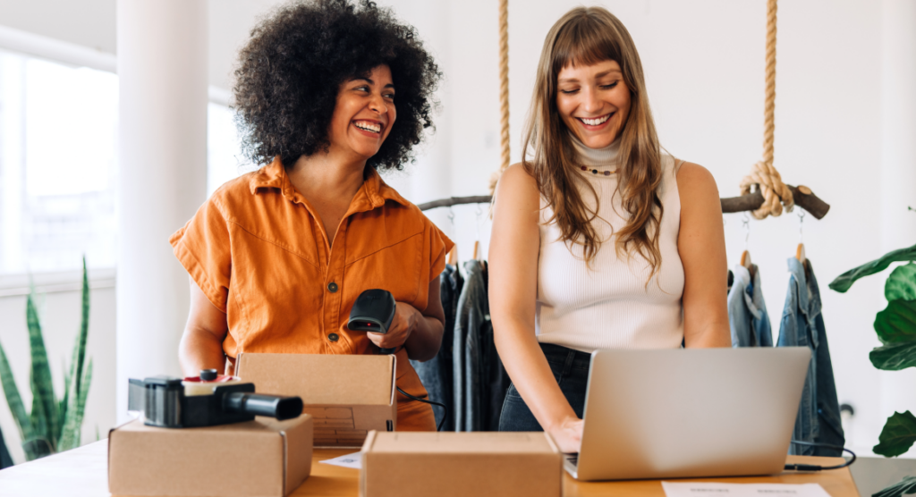 Two girls stand beside each other smiling as they work together to scan and package shipments.