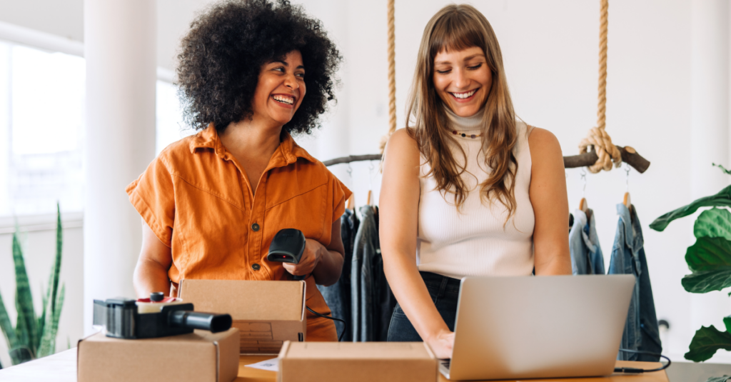 Two girls stand next to each other smiling as they work together to scan and package shipments.